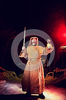 Medieval monk in canvas sackcloth robe with sword and lattern in dark forest and red light of moon on winter night. Fantasy or