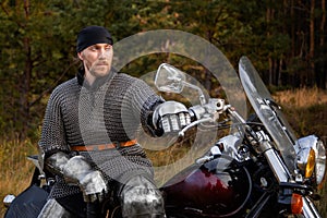 medieval warrior in armor sits on a motorcycle with a sword in his hands on the background of the forest