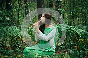 A medieval woman in a green dress plays on a wooden flute sitting in fern bushes. A girl in a gloomy forest plays music on a handm