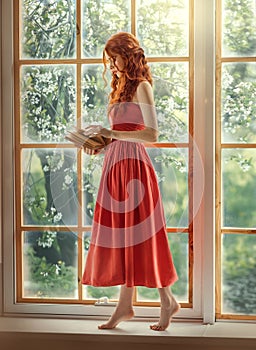 Medieval woman princess holds in hands reads book. Red-haired dreamlike girl stands on window barefoot, view summer