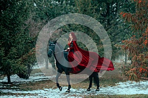Medieval woman princess in red dress sits astride black steed horse. Girl rider in vintage cloak cape train flies in