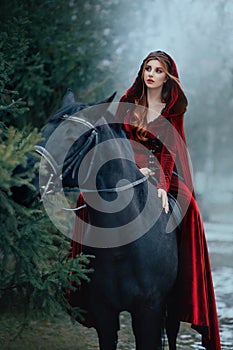 Medieval woman princess in red dress sits astride black steed horse. Girl rider in vintage cloak cape train flies in
