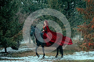 Medieval woman princess in red dress sits astride black steed horse. Girl rider in vintage cloak cape train flies in photo