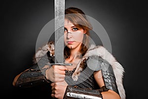 Medieval woman warrior in chain mail armor and polar fox fur on her shoulders standing with a sword in hand on a dark background.