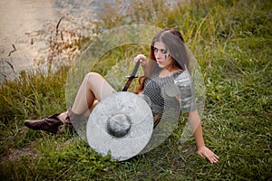 Medieval woman warrior in chain mail with a shield and a sword sits on the river bank in the evening