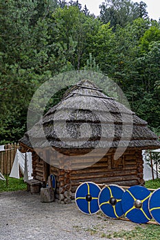 Medieval wooden house with a thatched roof, the military defense of an old warrior leaning on a float in the background of green