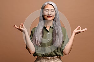 Meditation concept. Calm and satisfied mature woman holds her hands in zen gesture, keeps eyes closed, feels appeasement