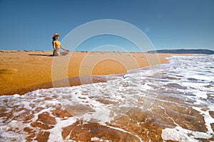 Meditation by the sea on a sunny day. Young caucasian woman in a yellow swimsuit sits on the golden sand in a lotus