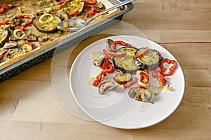 Mediterranean oven vegetables baked in olive oil with herbs and spices on a plate and on a baking tray, wooden table, vegetarian