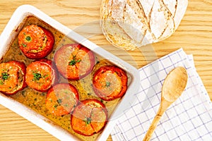 Mediterranean stuffed tomatoes with meat and bread crumbs,  in a white oven dish, aside a kitchen towel, and bread loaf on oak