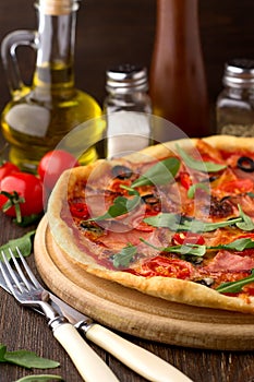 Mediterranean traditional pizza with ham, tomatoes, olives and herbs on wooden table.