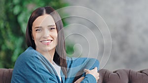 Medium close-up portrait of casual smiling young woman looking at camera in living room