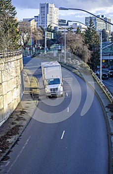 Medium duty rig white semi truck transporting goods in box trailer going to highway entrance running from the city street downhill