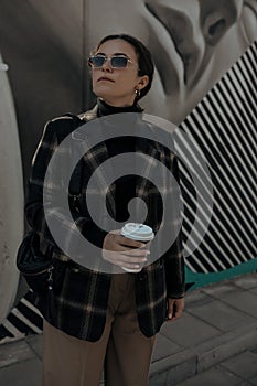 brunette woman in trendy clothes and in sunglasses with paper cup coffee cup in hand