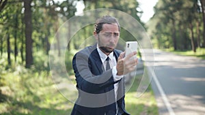 Medium shot of anxious male driver searching mobile network on suburban sunny road with broken vehicle at background