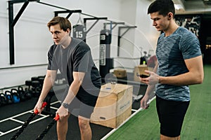 Medium shot of beginner sportsman hard time doing CrossFit workout with battle ropes by personal trainer standing with