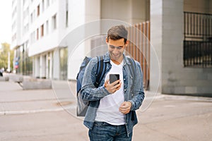 Medium shot of cheerful handsome male courier with thermo backpack using navigation app on phone standing in city street