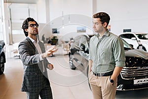 Medium shot of competent male car dealer in business suit having conversation with serious young man client at showroom