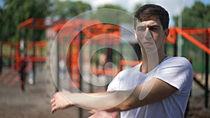 Medium shot of confident young sportsman stretching hand muscles in slow motion in sunshine outdoors. Serious Caucasian