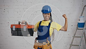 Medium shot of a dark-skinned young female construction worker standing in the room, wearing a tool belt, raising a tool