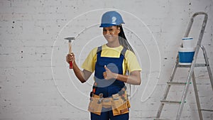 Medium shot of a dark-skinned young female construction worker standing in the room, wearing a tool belt, showing a