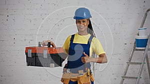 Medium shot of a dark-skinned young female construction worker standing in the room, wearing a tool belt, showing a tool