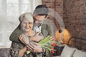 medium shot of elderly happy grandma with her middle-aged grandson holding flowers and celebrating grandparent& x27;s day