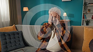 Medium shot of an eldery, retired man, senior citizen sitting on a couch, massaging his temples and head due to a severe