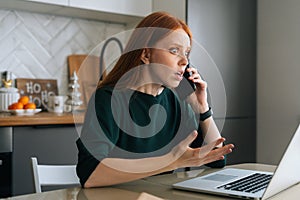 Medium shot of furious young woman talking on mobile phone and using laptop sitting at table in kitchen with modern