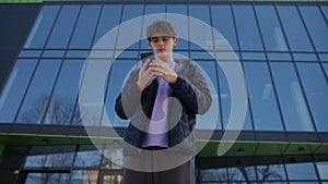 Medium shot of handsome young man working on smartphone stands on the street in front of a modern building, then looks