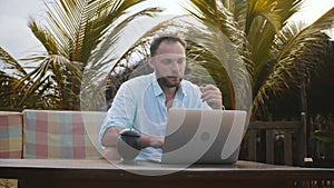 Medium shot of happy successful young creative worker man using laptop to work online under palm trees at exotic beach