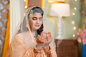 Medium shot of Indian Muslim woman praying or namaz with eyes closed during Ramadan feast celebration at home - concept