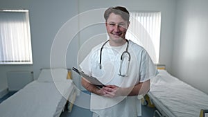 Medium shot of intelligent positive Caucasian male doctor standing in hospital ward with medical history smiling looking