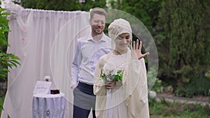 Medium shot of joyful young Middle Eastern bride showing off with ring on finger and wedding bouquet as Caucasian groom