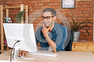 Medium shot of laughing young man wearing stylish glasses working on desktop computer sitting at desk at home office in