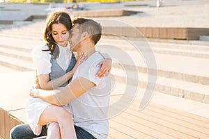 Medium shot portrait of beautiful young couple resting in city park sitting hugging on bench in summer sunny day.