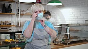 Medium shot portrait of Caucasian woman putting on face mask looking at camera standing in coffee shop. Blurred man