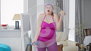 Medium shot portrait of charming obese woman lifting dumbbell eating unhealthy fast food. Overweight Caucasian young