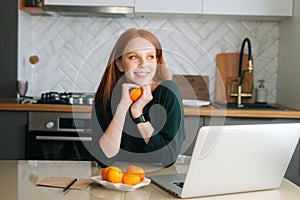 Medium shot portrait of cheerful redhead young woman sitting at table with laptop and holding tangerine in hand