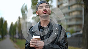 Medium shot portrait of confident gay man drinking coffee from paper cup standing outdoors on city street. Handsome