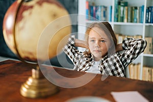 Medium shot portrait of curious child girl sitting on chair with hands behind head and looking at little globe on table