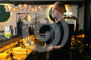Medium shot portrait of handsome young barman standing near bar counter waiting for customers orders on blurred