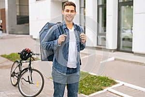 Medium shot portrait of happy handsome young delivery man with large thermo backpack standing on city street.