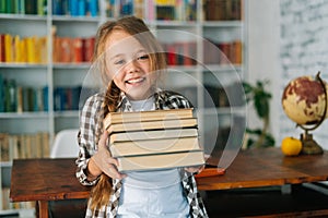 Medium shot portrait of joyful elementary child school girl holding stack of books in library at school, looking at