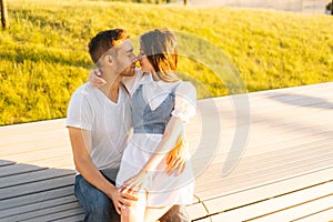 Medium shot portrait of loving young couple hugging and kissing sitting on bench in city park in summer sunny day.