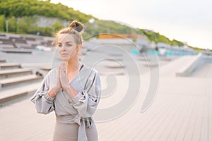 Medium shot portrait of meditative Caucasian young woman practicing yoga performing namaste pose outside in city park.