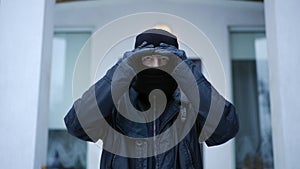 Medium shot portrait of serious male burglar putting on ski mask turning walking to empty house on winter day