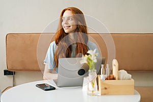 Medium shot portrait of smiling young woman working typing using on laptop computer sitting at table in cozy cafe