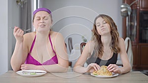 Medium shot portrait of young obese woman eating healthful organic cucumber as slim beautiful millennial enjoying