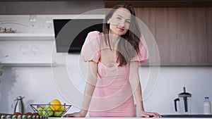 Medium shot portrait of young slim beautiful brunette woman with grey eyes looking at camera smiling standing at kitchen
